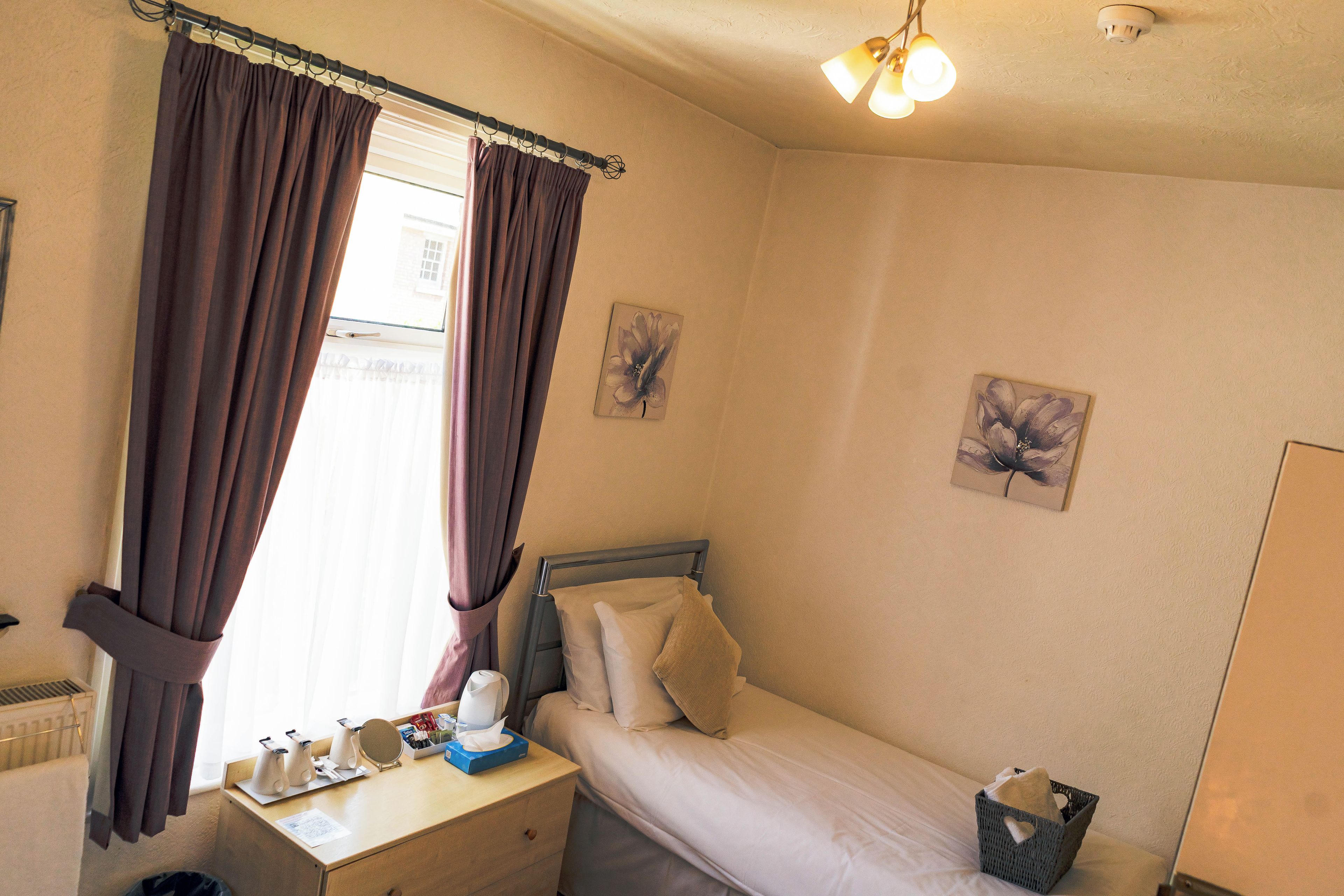 A small, neatly arranged bedroom with a single bed, a bedside table with a lamp, a window with curtains, and two wall-mounted flower paintings.