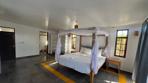 A bedroom with a four-poster bed, a wooden nightstand, and a window with a view of greenery. -  2Bdr zen villa private pool + cleaning (Diani Beach)