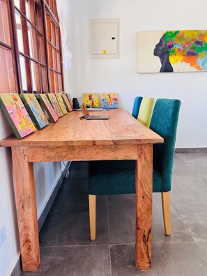 Ein Holztisch mit farbenfrohen Bildern links, darauf ein Laptop und ein Stuhl mit einem grünen Kissen.