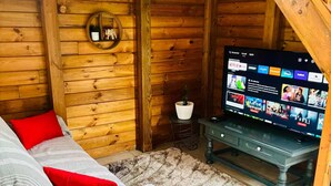 Interior acolhedor de uma cabana de madeira, com parede rústica, sofá com almofadas vermelhas, televisão exibindo um serviço de streaming e uma pequena planta em vaso.