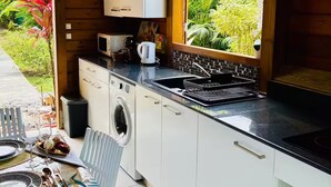 Cozinha compacta com armários brancos, micro-ondas, chaleira e pia. Há uma janela com vista para áreas verdes.
