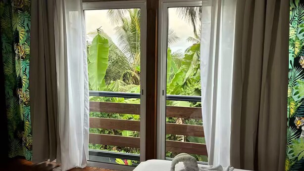 Quarto com cama, cortinas e vista para a vegetação tropical através de uma porta de correr.