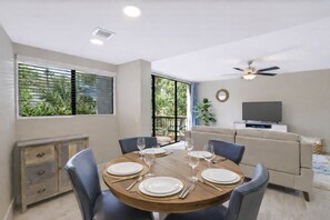 A dining area with a round table set for four, a sofa, a television, and a ceiling fan. - Poolside Villa, Walk to Beach - Coligny Hotspots Close (Hilton Head Island)