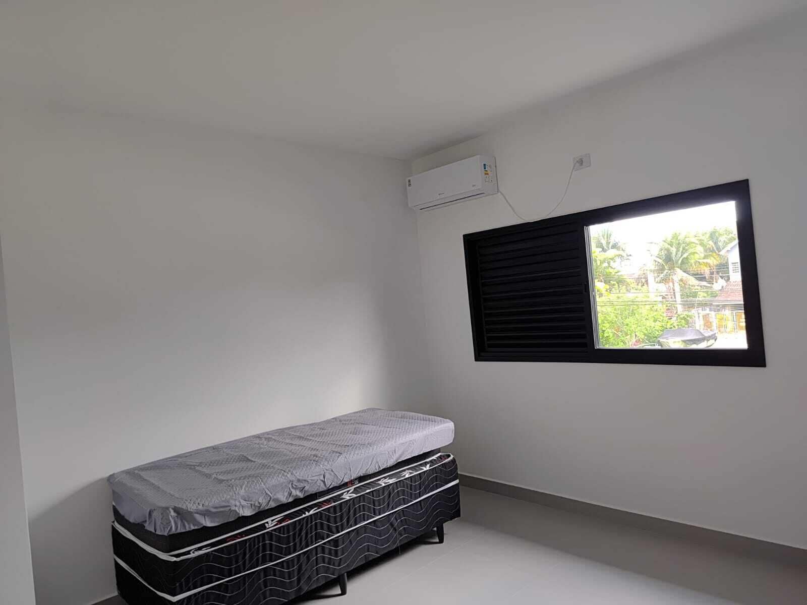 Una habitación pequeña y escasamente amoblada, con una cama individual, una ventana con persianas negras y un equipo de aire acondicionado en la pared.