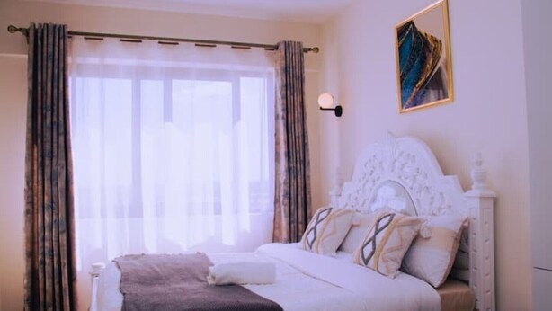 Ein Schlafzimmer mit einem großen Bett, einem Kopfteil, einem Nachttisch, Vorhängen an einem Fenster und einer Wandlampe.