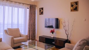 Ein Wohnzimmer mit einer Couch, einem Glastisch, einem Flachbildfernseher, einer dunklen Holzkommode und einer Blumenvase.