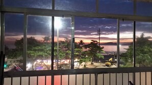 Blick aus einem Fenster auf einen Sonnenuntergang über einem Gewässer, mit Bäumen und einer beleuchteten Straße im Vordergrund.