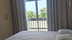 Ein Schlafzimmer mit Bett, Deckenventilator, Fenster mit Vorhängen und Blick ins Freie.