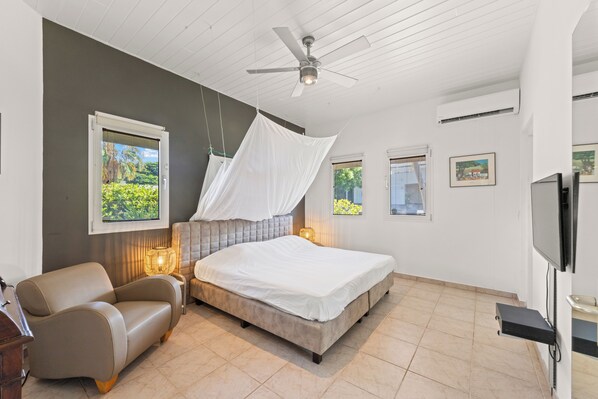 A bedroom with a bed, a ceiling fan, a chair, a TV, and a window with a view of greenery. - Beautiful Oceanview Home with private pool, In walking distance from Mambo Beach (Willemstad)