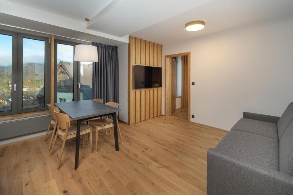 Un salon moderne comprenant un coin repas, un canapé gris, un plancher en bois et un téléviseur fixé au mur.