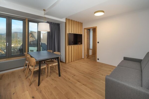 Un salon moderne comprenant un coin repas, un canapé gris, un plancher en bois et un téléviseur fixé au mur.