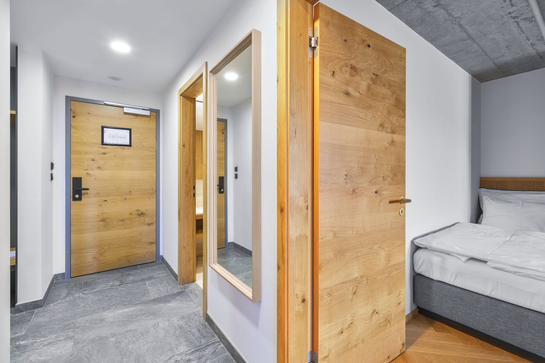 A modern hotel room with a wooden door, a large mirror, and a bed with white linens.