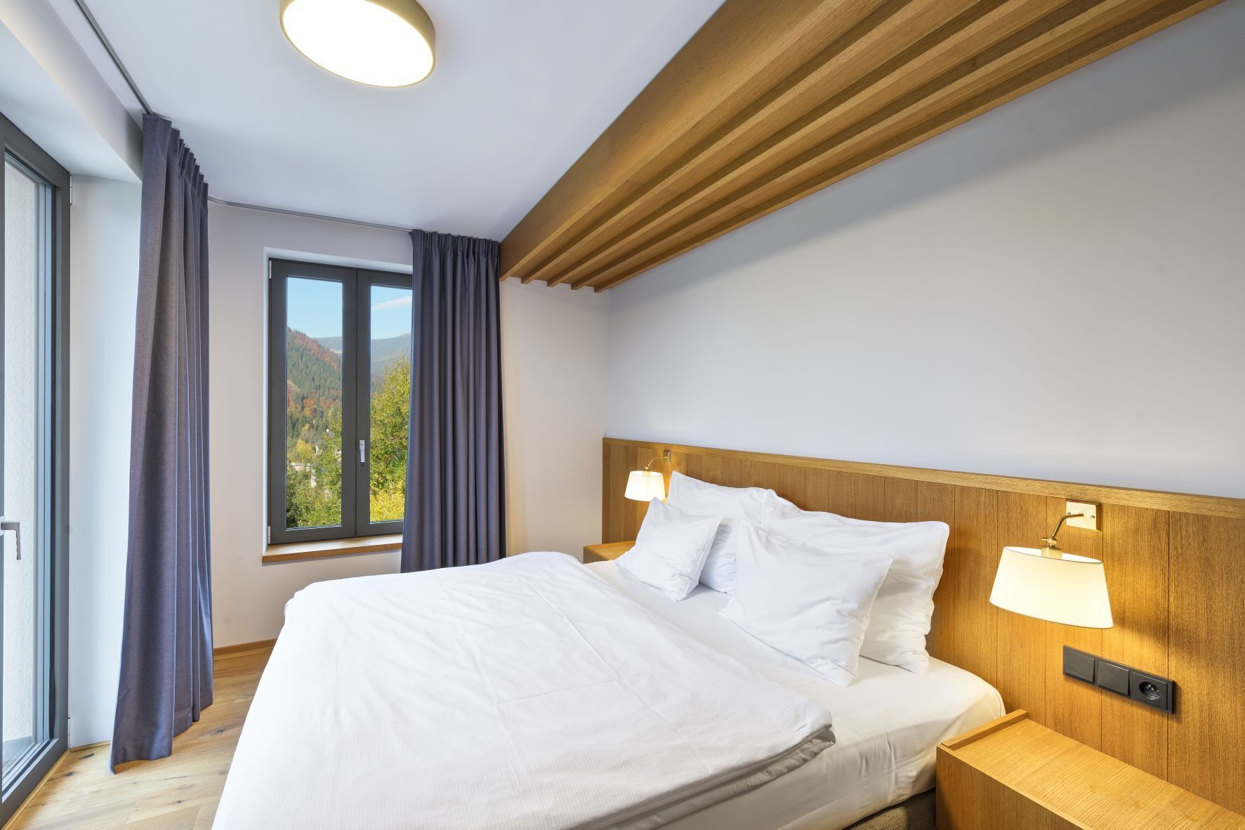 Una habitación de hotel moderna con una cama grande, cabecera de madera, dos lámparas de noche y un ventanal con cortinas.