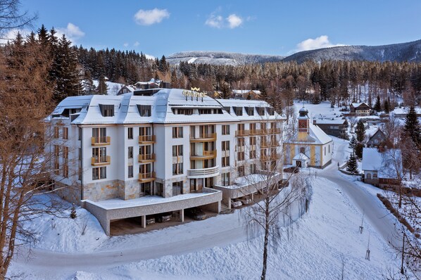 Ein mehrstöckiges Hotel mit schneebedecktem Dach, einem markanten Turm und Blick auf ein verschneites Gebirge.