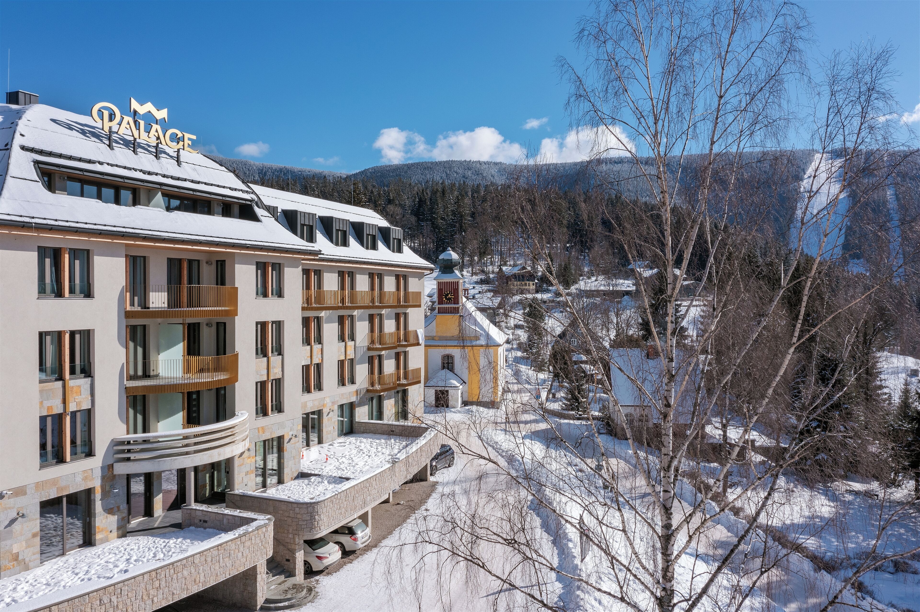Un hôtel appelé « Palace », doté d’un design moderne, de nombreux balcons et offrant une vue sur un paysage enneigé avec des montagnes en arrière-plan.