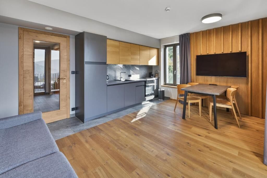 A modern kitchen with grey cabinets, a wooden dining table, and a flat-screen TV mounted on the wall.