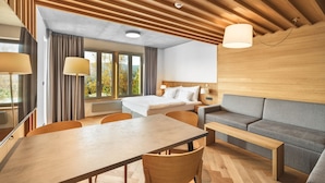 Ein modernes Hotelzimmer mit Holzboden, einem Bett, einer Couch, einem Esstisch und Blick auf Bäume durch große Fenster.