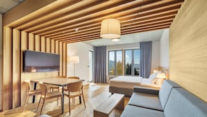 Ein modernes Hotelzimmer mit Holzboden, einem Flachbildfernseher, einer Essecke mit Tisch und Stühlen, einem Sofa und einem großen Fenster mit Blick ins Freie.