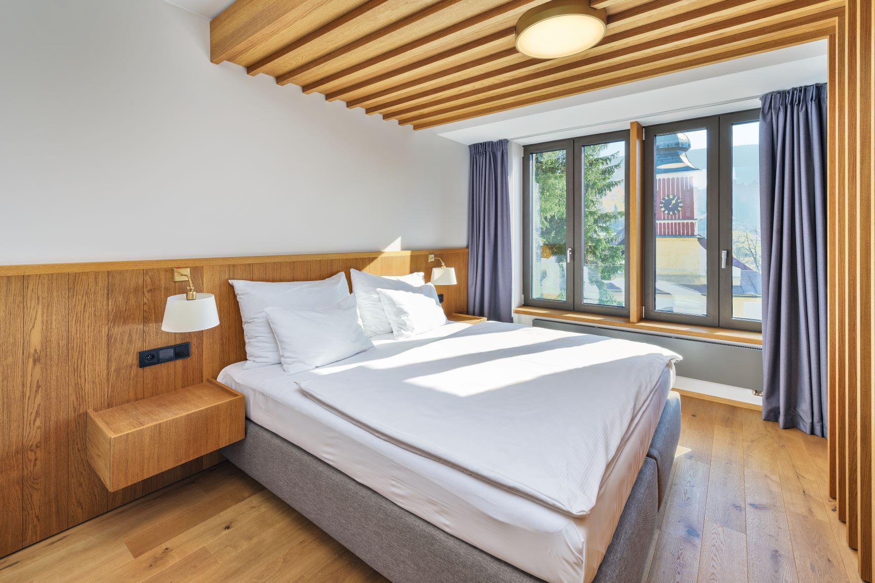 Un dormitorio moderno con cabecera de madera, una cama grande con ropa blanca, una mesita de noche con lámpara y una ventana con cortinas.