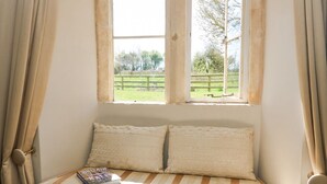 A cozy reading nook with a cushioned bench, a window with a view of a green landscape, and a book titled "BRITAIN". - The Smithy (Moreton-in-Marsh)