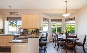 A kitchen with wooden cabinets, a granite countertop, a dining area with a table and chairs, and a view of greenery outside. - Gated Rancho Mirage Oasis w/ Pool & Spa (Rancho Mirage)