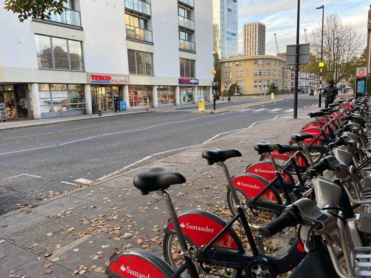 En række Santander cykler parkeret langs et fortov ved siden af en bygning med en Tesco Express butik.