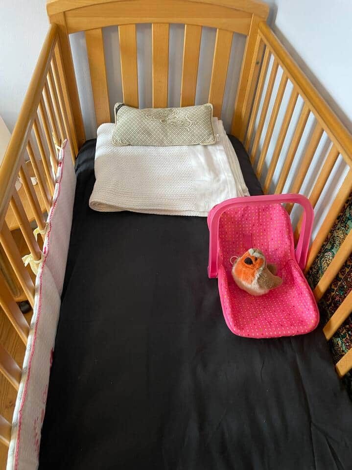 Un berceau en bois avec un matelas noir, une couverture blanche, un oreiller vert et un panier à jouets rose contenant un canard en plastique.