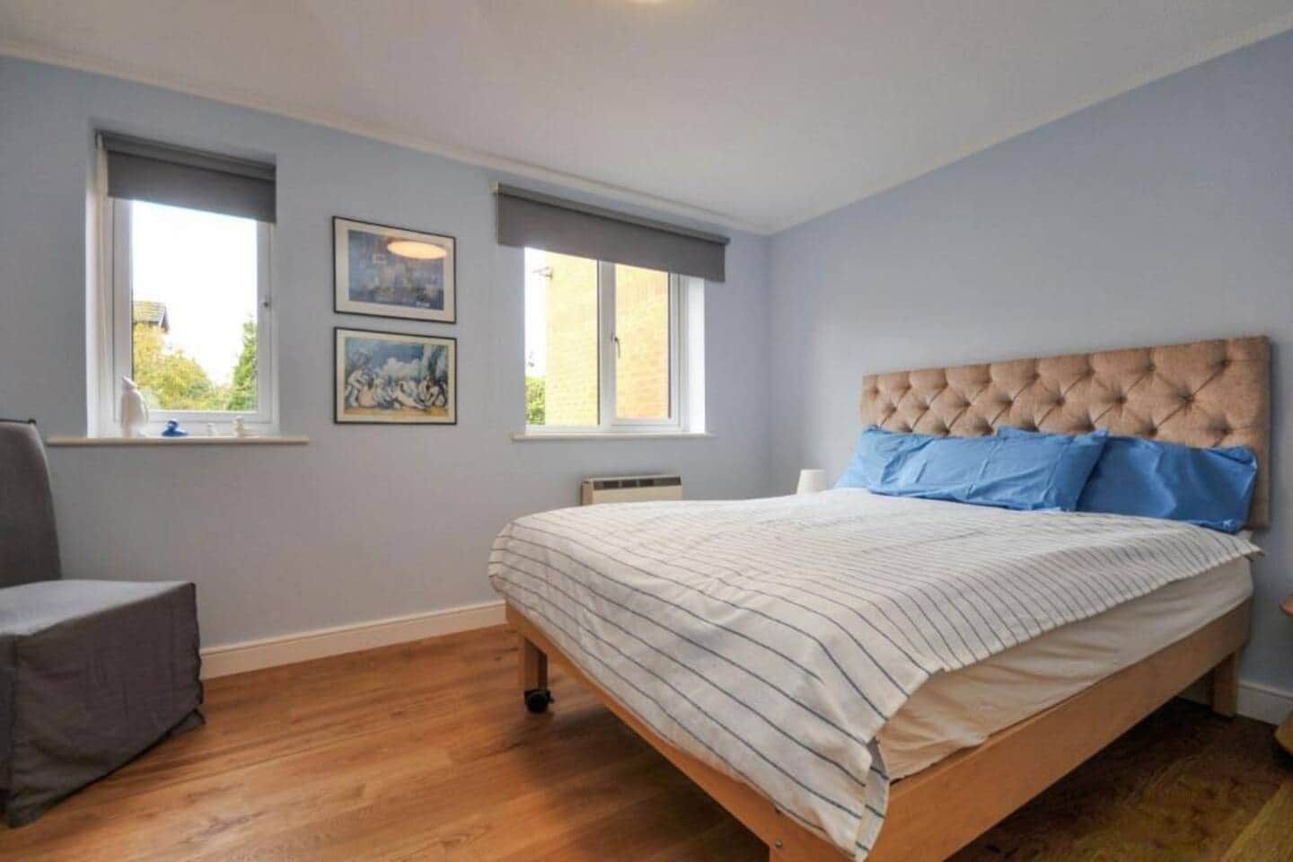 Une chambre à coucher avec un lit, deux fenêtres, une chaise et des œuvres d’art accrochées au mur.
