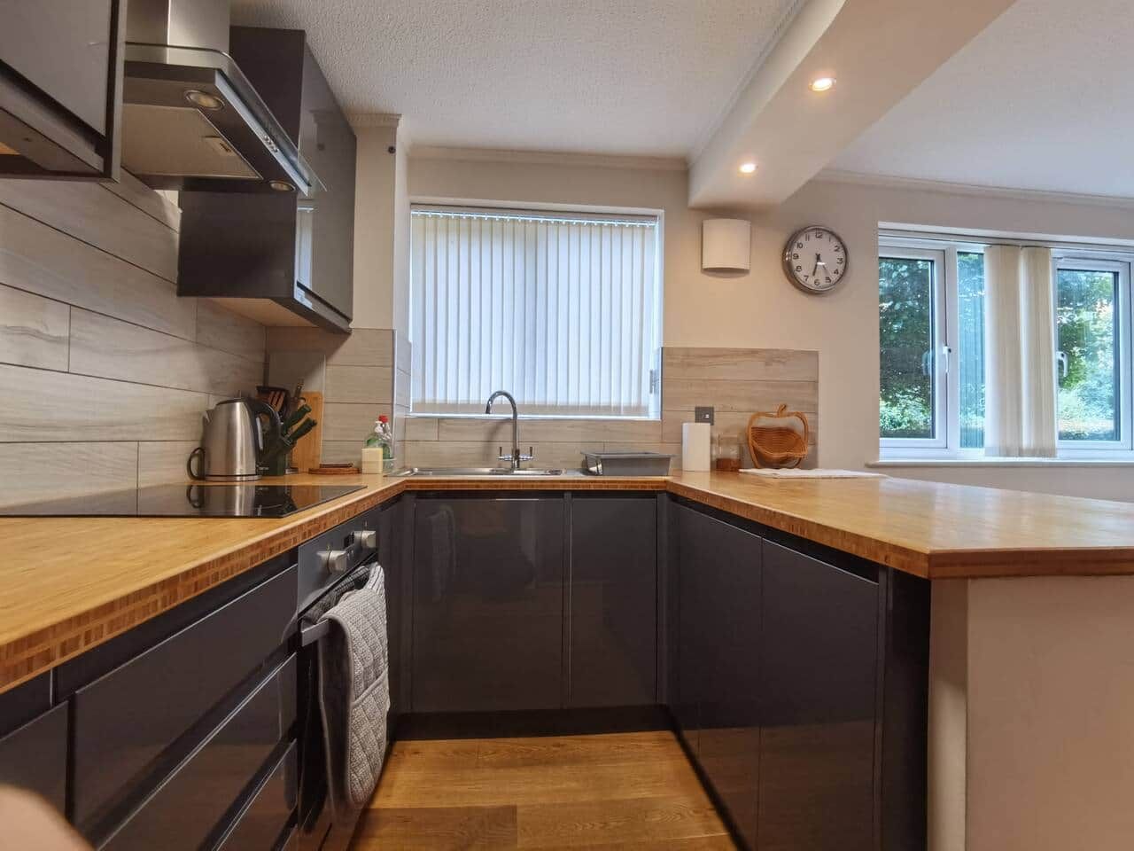 Une cuisine moderne avec des meubles de couleur sombre, un plan de travail en bois, une bouilloire en acier inoxydable et une fenêtre équipée de stores.