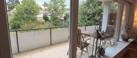 Un balcón con vistas a un jardín y edificios, una mesa de madera con sillas y una barandilla.