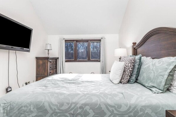 Une chambre à coucher avec un lit, un téléviseur fixé au mur, une fenêtre avec des rideaux et une tête de lit en bois.