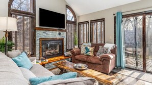 A spacious living room with a fireplace, a large flat-screen TV, a brown leather sofa, and a wooden coffee table. - Luxury, Golf-Themed Townhome (Galena)