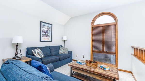 A living room with a blue sofa, a wooden coffee table, and a framed picture on the wall. - 3BR, 3BA Townhome w/ Sunporch + Fireplace (Galena)