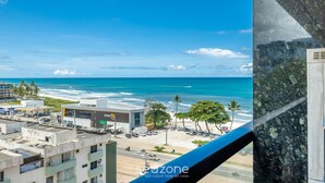 A coastal cityscape with a view of the ocean, a parking lot, and a building with a 'Fit' sign. - Apto Vista Mar Lateral em Frente a Praia BDT0608 (Ilhéus)