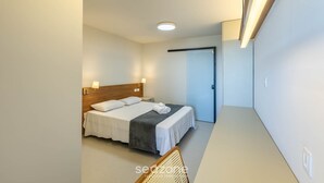 A modern hotel room with a neatly made bed, a wooden headboard, and a bedside table with a lamp. - Apto Vista Mar Lateral em Frente a Praia BDT0608 (Ilhéus)