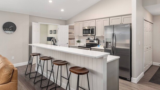 A modern kitchen with white cabinetry, a stainless steel refrigerator, a microwave, and a built-in oven. There are three stools at the bar counter. - The Hobby Horse - 8 miles from WEC ! (Ocala)