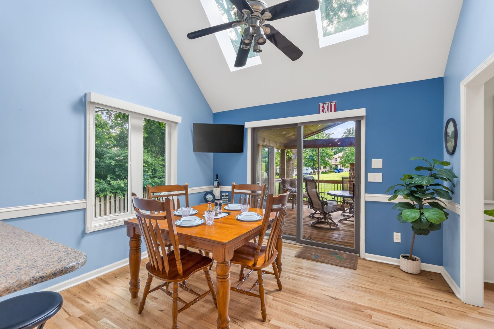 A dining area with a wooden table set for four, a ceiling fan, and a view of the outdoors through a glass door.