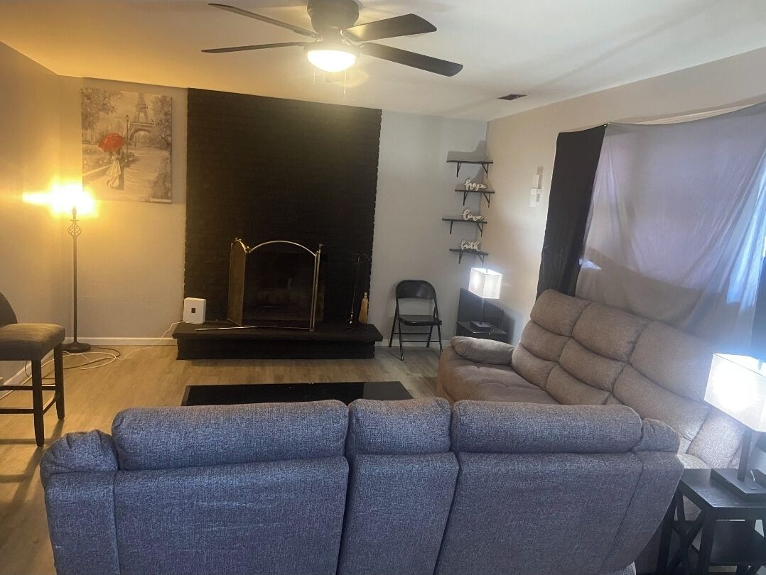 A living room with a sofa, a fireplace, a ceiling fan, and a wall-mounted shelf with decorative items.