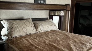 Uma cama com um edredom estampado e almofadas, uma cabeceira com uma imagem, e uma janela com cortinas.