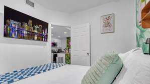 Une chambre avec un sol à carreaux, un lit avec un oreiller vert, un tableau représentant un paysage urbain accroché au mur, et une cuisine en arrière-plan.