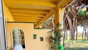 A covered outdoor seating area with a sofa, a table, and a lamp. There are plants and a view of a garden. - ✨ Luminous townhouse overlooking the Golf on the beach of Novo Sancti Petri (Chiclana de la Frontera)