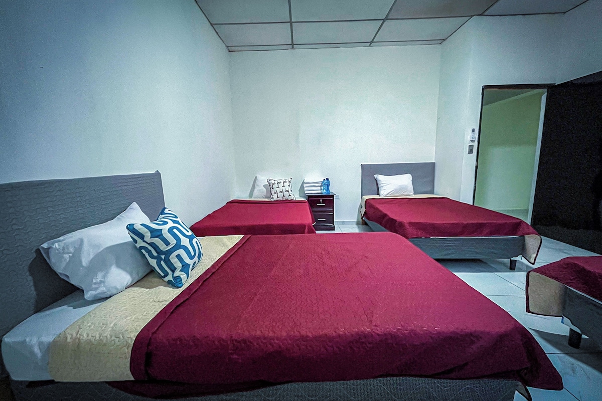 A room with four beds, each with a red bedspread and white pillows. There is a small table with a water bottle and a white pillow with text. The room has white walls and a tiled floor.