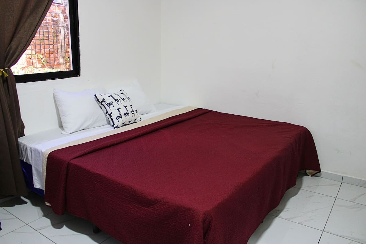 A single bed with a red bedspread, white pillows, and a window with a view of a brick wall.
