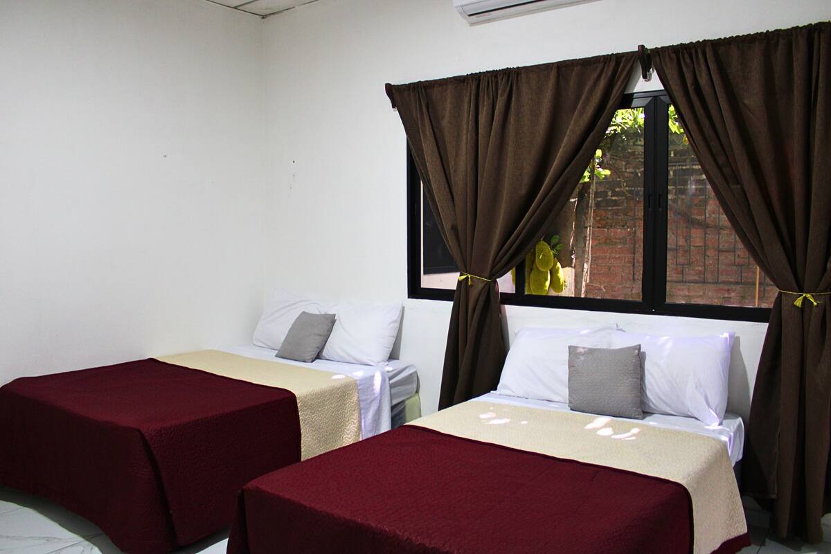 A room with two beds, each with a red bedspread and white linens, and a window with brown curtains.