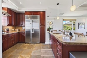 A modern kitchen with a stainless steel refrigerator, wooden cabinets, granite countertops, and a large island with a sink. - Spacious Ocean View condo with AC in Mauna Kea Resort (Waimea)
