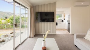 A modern living room with a large sliding glass door leading to a balcony, a flat-screen TV mounted on the wall, and a minimalist kitchen in the background.