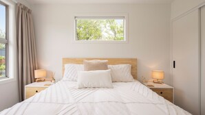 A neatly made bed with white bedding, two bedside tables with lamps, a window with curtains, and a view of trees outside.