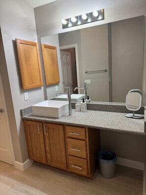 A bathroom with a double sink, wooden cabinets, a vanity mirror, and a trash can. - Beautiful Courtyard Villa near Lake Sumter Landing (The Villages)