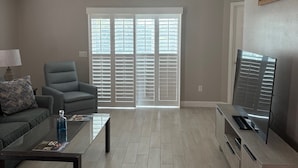 A living room with a ceiling fan, a sofa, a coffee table, a TV stand, and a large window with blinds. - Beautiful Courtyard Villa near Lake Sumter Landing (The Villages)