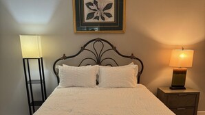 A neatly made bed with a white duvet, a framed floral artwork above it, and two bedside lamps on a nightstand. - Beautiful Courtyard Villa near Lake Sumter Landing (The Villages)